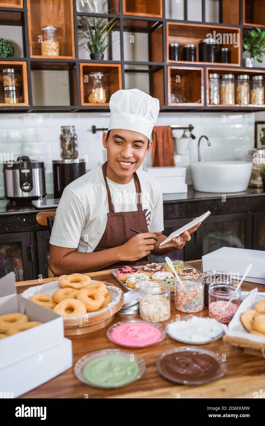 Uomo asiatico che indossa cappello e grembiule da chef sorride come lui  guarda le ciambelle sul tavolo mentre scrivi un notebook mentre si è seduti  in cucina Foto stock - Alamy
