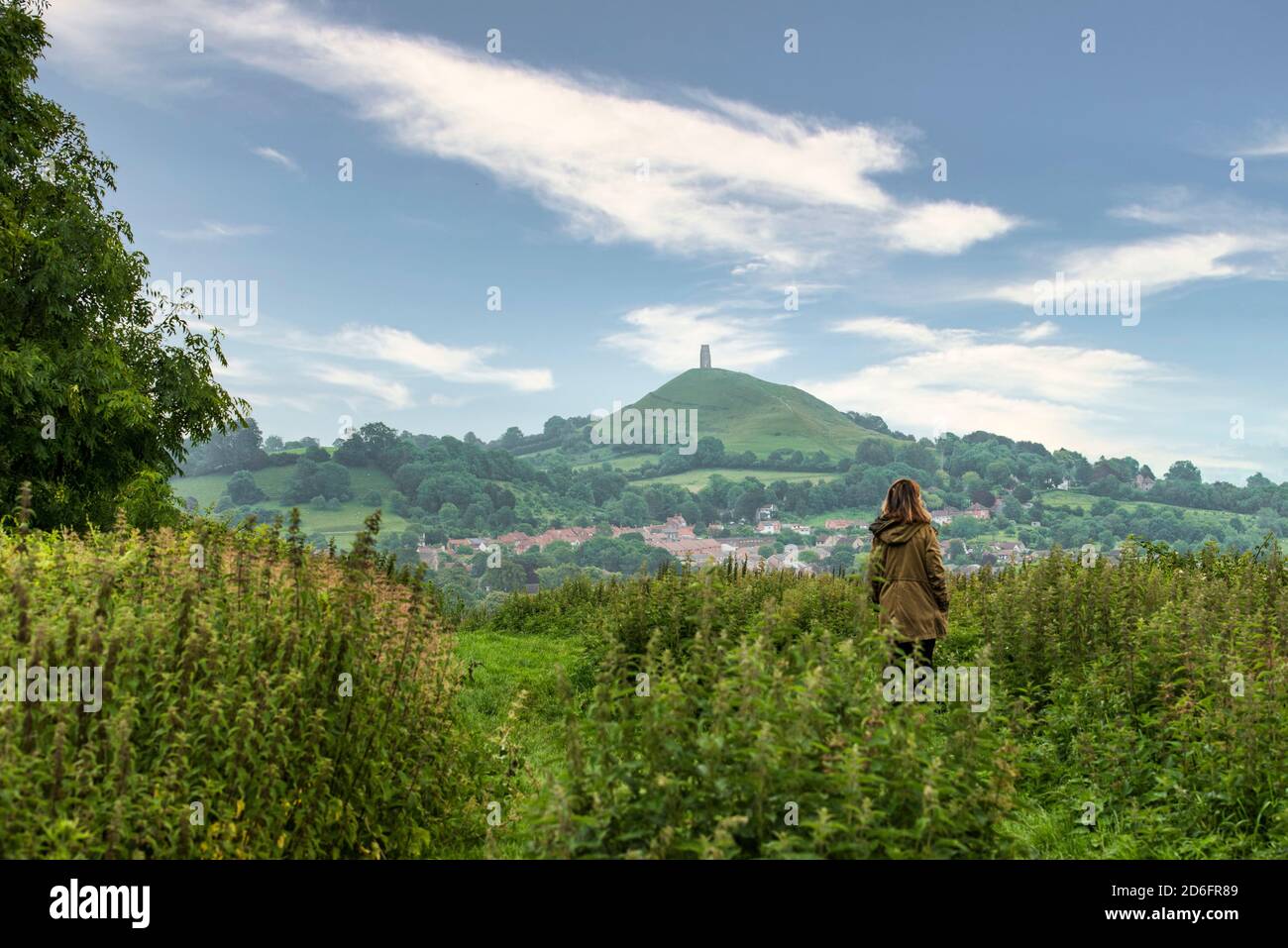 Lone femmina contemplando Glastonbury Tor in lontananza. In piedi in un campo locale lontano dai turisti. Cielo blu con un sacco di spazio per la copia. Foto Stock