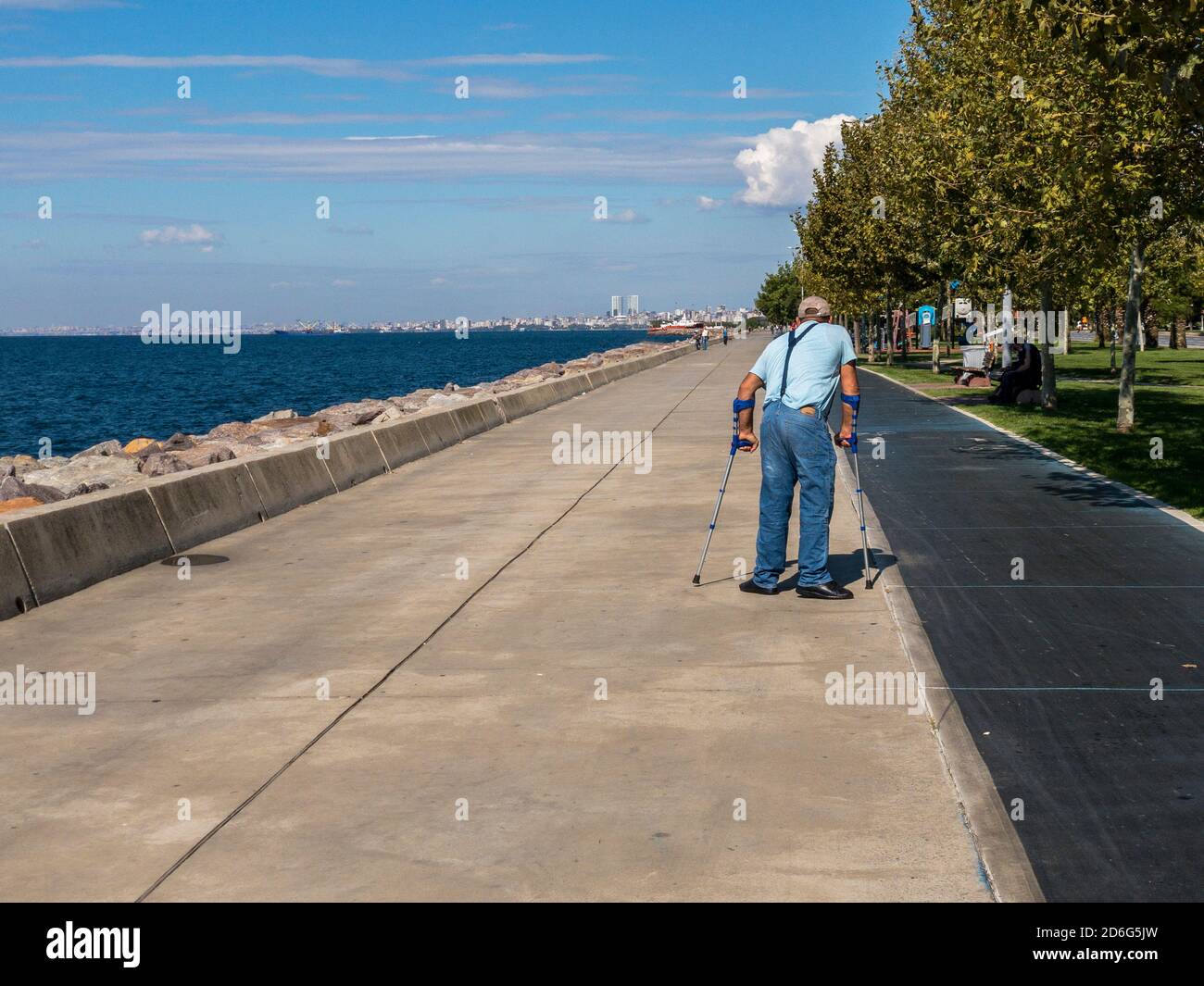 Istanbul / Turchia - 10.1.2020; un anziano cammina con le grondole su un marciapiede di parco circondato da alberi vicino al mare in una giornata di sole. Foto Stock