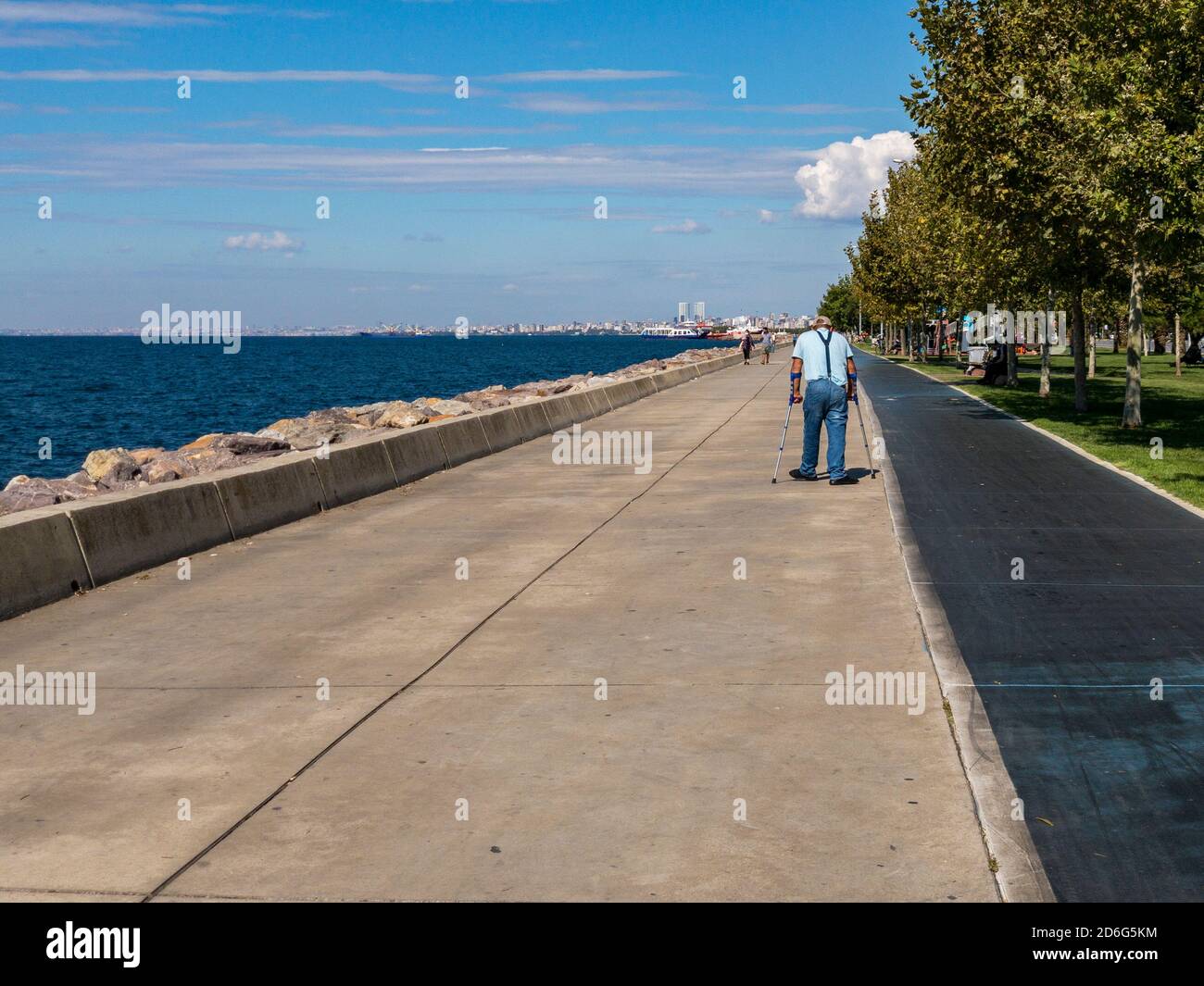 Istanbul / Turchia - 10.1.2020; un anziano cammina con le grondole su un marciapiede di parco circondato da alberi vicino al mare in una giornata di sole. Foto Stock