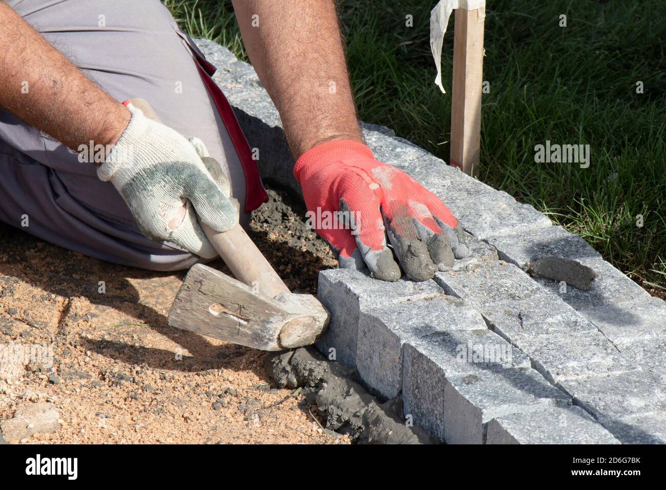 Mani in guanti di un lavoratore costruttore che posa sul granito ciottolato sett su sabbia e Malta con martello, marciapiede pavimentazione Foto Stock
