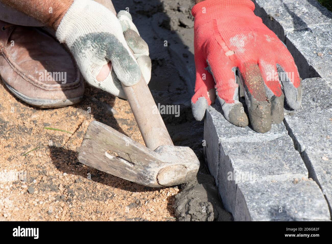 Mani in guanti di un lavoratore costruttore che posa sul granito ciottolato sett su sabbia e Malta con martello, marciapiede pavimentazione sul cantiere Foto Stock