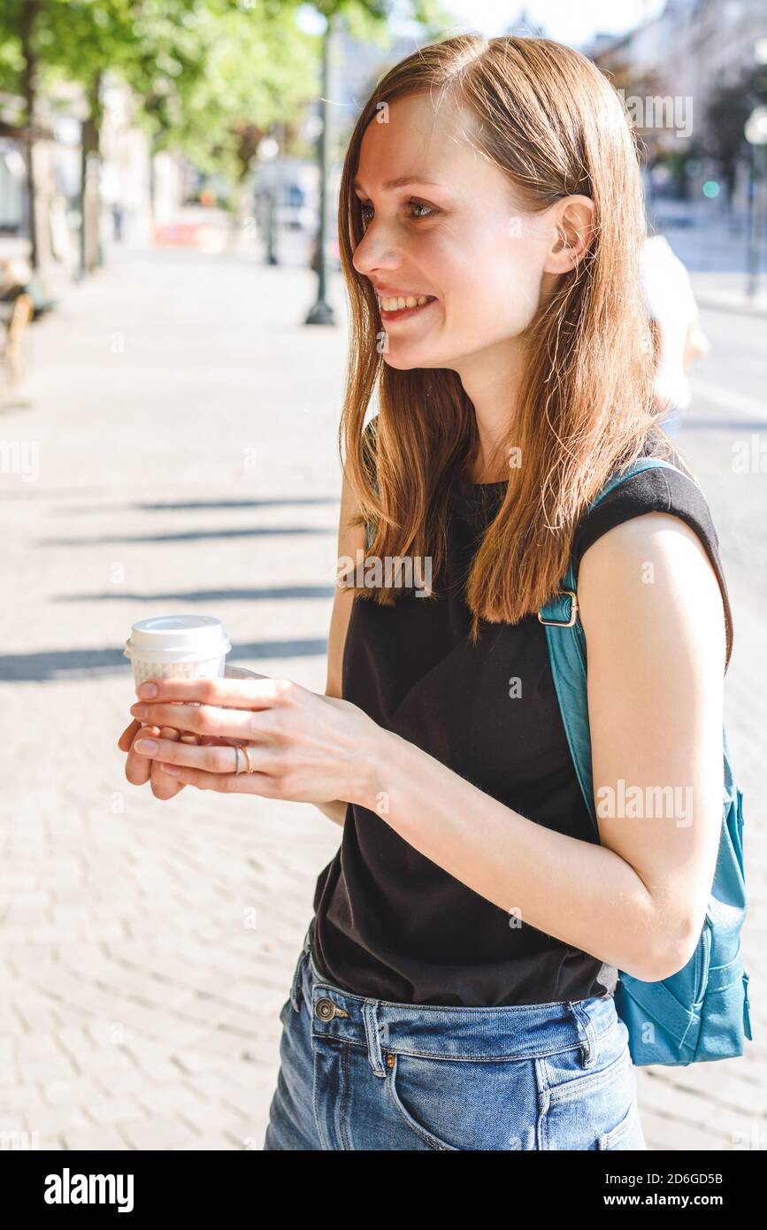 ritratto verticale di una giovane bella donna caucasica rossa in piedi in strada con una tazza di caffè da portare via e sorridendo Foto Stock