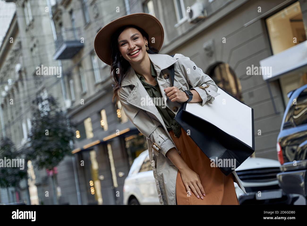 Giornata dello shopping. Giovane bella donna entusiasta in cappello con borsa per lo shopping guardando la macchina fotografica e sorridendo mentre si trova in piedi sulla strada della città, visitando le boutique. Moda, stile di vita della gente Foto Stock