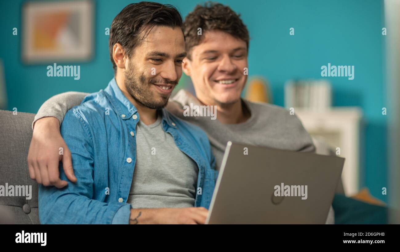 Adorabile coppia gay maschile trascorrere il tempo a casa. Si siedono su un divano e usano il laptop. Essi guardare divertenti video online. Il partner gli mette la mano intorno Foto Stock