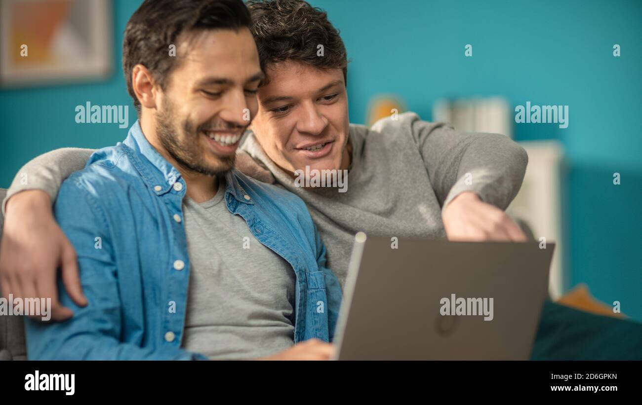 Adorabile coppia gay maschile trascorrere il tempo a casa. Si siedono su un divano e usano il laptop. Essi guardare divertenti video online. Il partner gli mette la mano intorno Foto Stock
