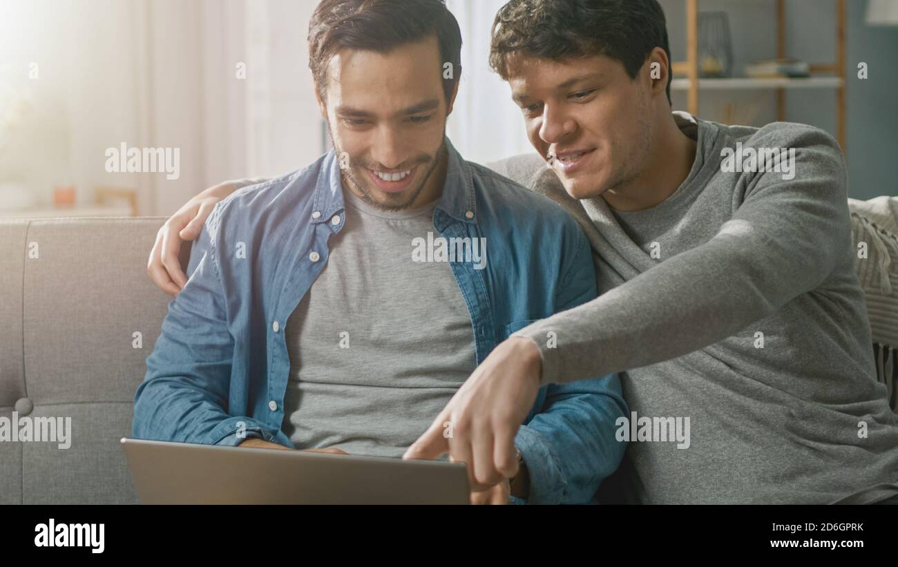 Adorabile coppia gay maschile trascorrere il tempo a casa. Si siedono su un divano e usano il laptop. Navigano online. Il compagno mette la mano intorno al suo amante. Camera Foto Stock