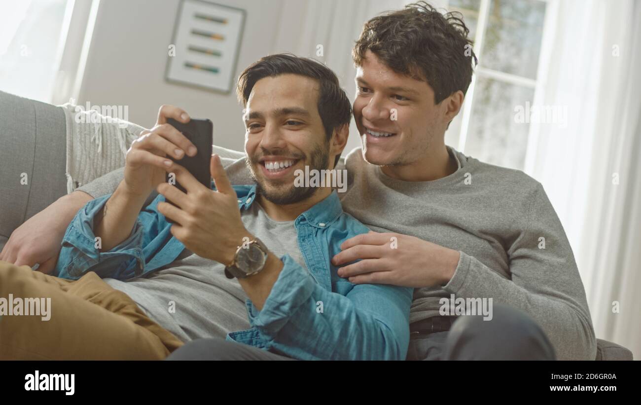 Carina coppia gay maschile trascorrere il tempo a casa. Sono sdraiati su un divano e utilizzano uno smartphone. Navigano online. La mano del compagno è intorno al suo amante Foto Stock