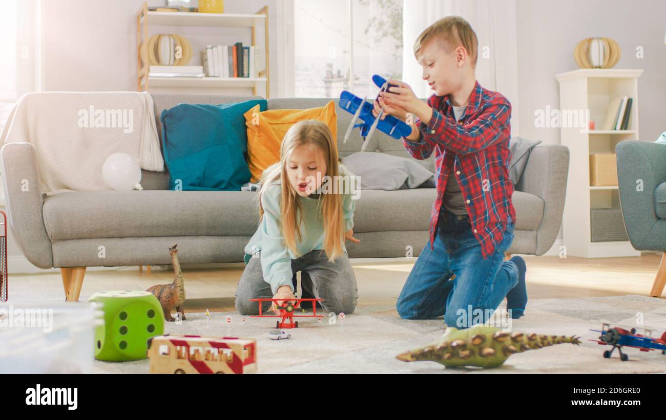 Nel salotto: Ragazzo e ragazza che giocano con gli aeroplani del giocattolo e i dinosauri mentre si siedono su un tappeto. Soggiorno soleggiato con bambini che si divertano. Foto Stock
