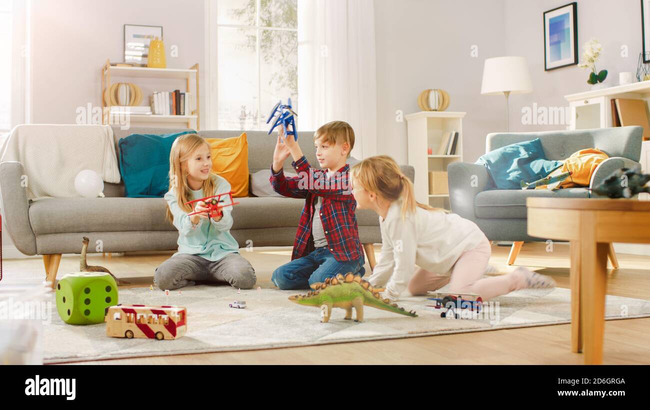 Nel salotto: Ragazzo e ragazza che giocano con gli aeroplani del giocattolo e i dinosauri mentre si siedono su un tappeto. Soggiorno soleggiato con bambini che si divertano. Foto Stock