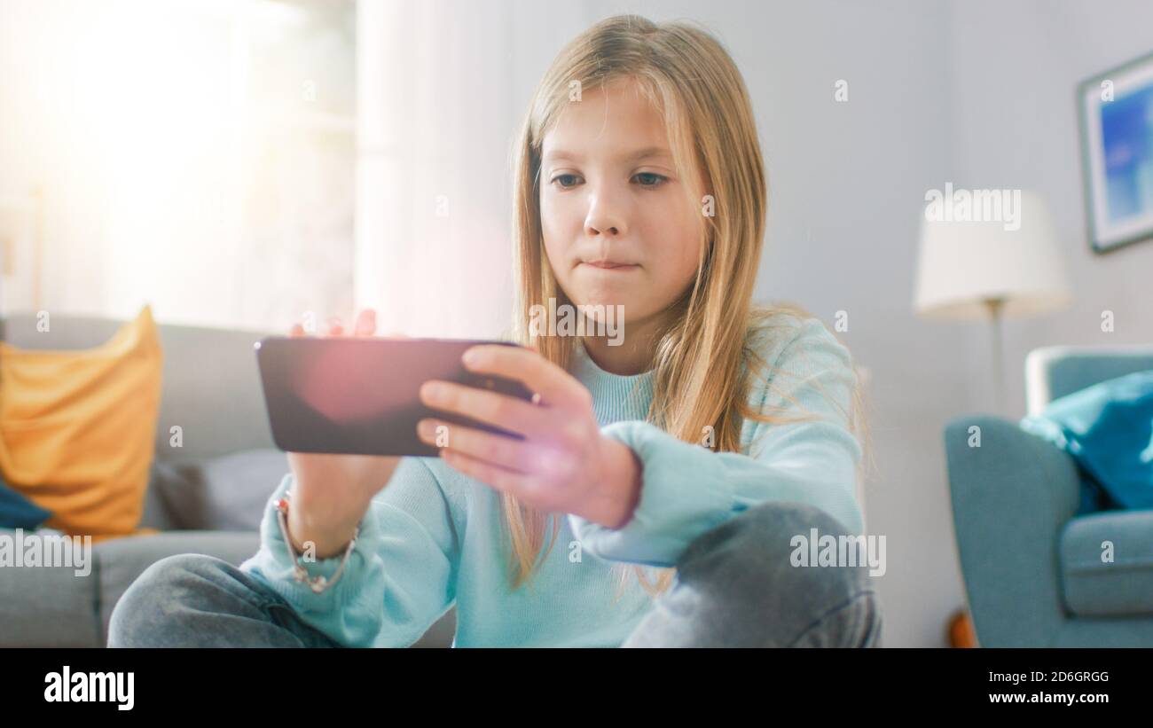 Ritratto scatto di una ragazza intelligente carino seduto su un tappeto giocando in videogioco sul suo smartphone, tiene e utilizza il cellulare in orizzontale Foto Stock
