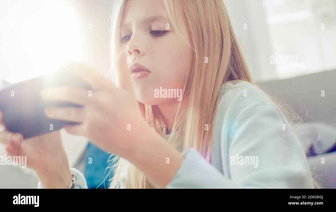 Primo piano Ritratto di una ragazza intelligente seduta su un tappeto a casa giocando in videogioco sul suo smartphone, tiene e utilizza il cellulare in orizzontale Foto Stock