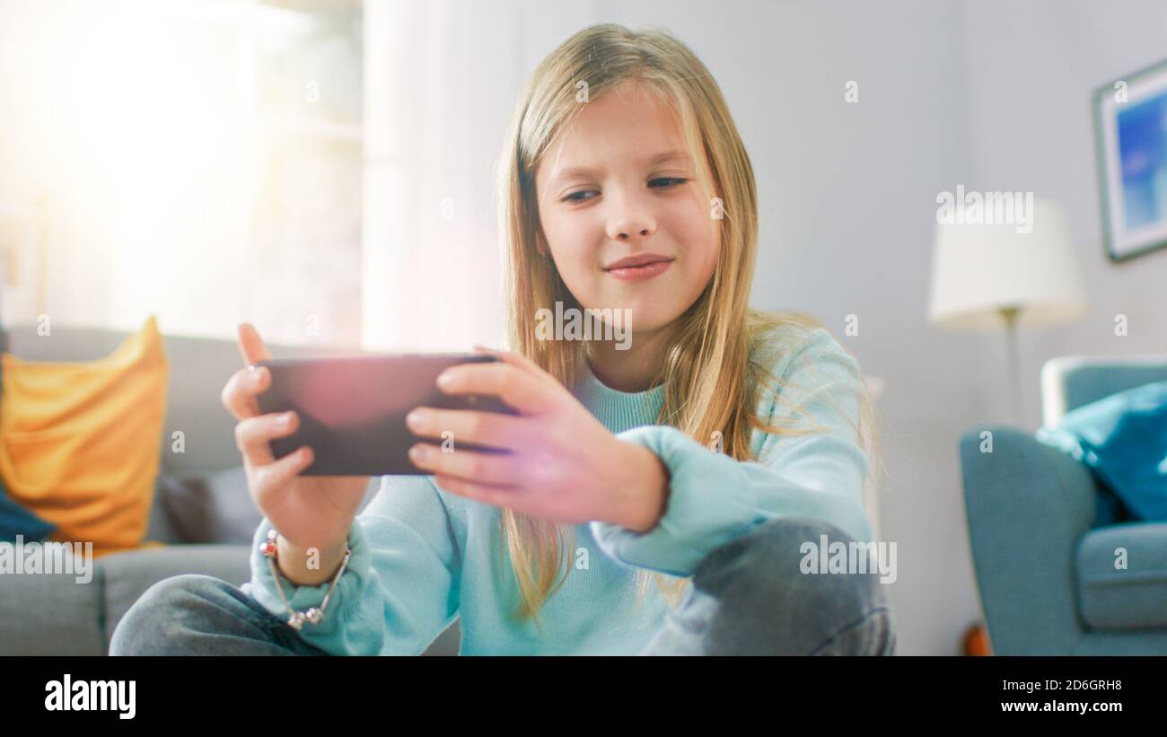 Ritratto scatto di una ragazza intelligente carino seduto su un tappeto giocando in videogioco sul suo smartphone, tiene e utilizza il cellulare in orizzontale Foto Stock