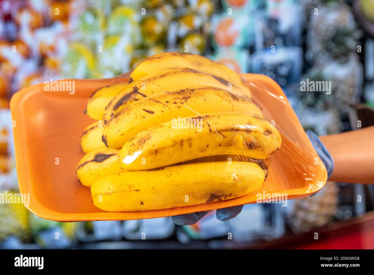 Nuova normalità dopo la pandemia di Covid-19: Una mano con guanti di protezione blu tiene una piastra di plastica avvolta con banane. In verdure di sfondo, frutta Foto Stock