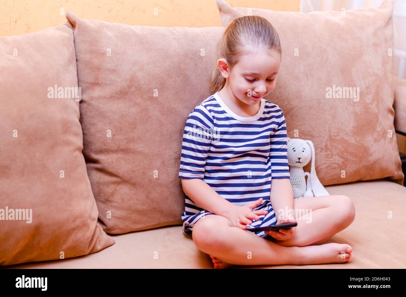 La bambina carina si siede su un divano a casa e tiene un telefono cellulare in mano. La comunicazione remota su un telefono cellulare registra i video di famiglia sul phon Foto Stock