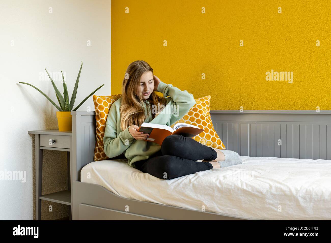 Giovane ragazza bionda teenage che legge un libro a casa dentro parte anteriore di una parete gialla Foto Stock