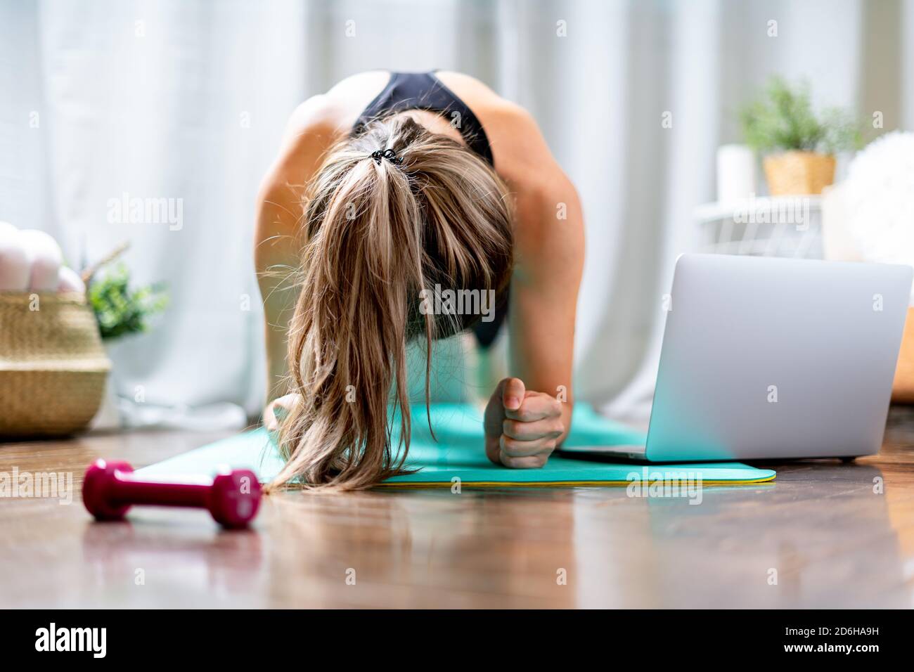 Giovane donna fitness che lavora fuori e usando il laptop e i manubri a casa in soggiorno, facendo yoga o pilates esercizio su tappeto turchese, in piedi in pl Foto Stock