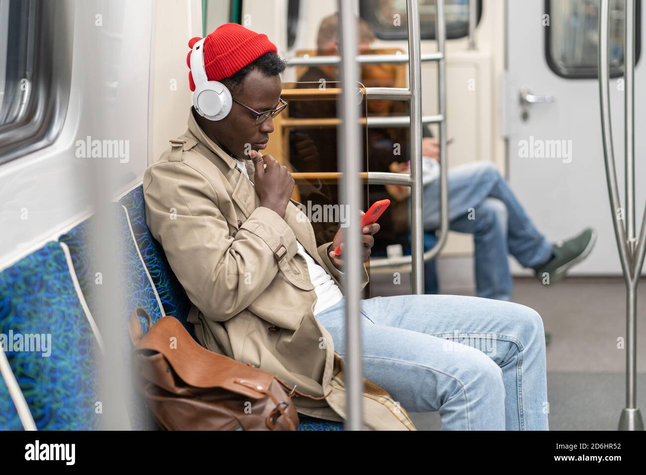 Uomo afro-americano in cappello rosso, trench cappotto siting in treno della metropolitana, utilizzando il telefono cellulare, ascolta la musica con le cuffie wireless in tr pubblico Foto Stock