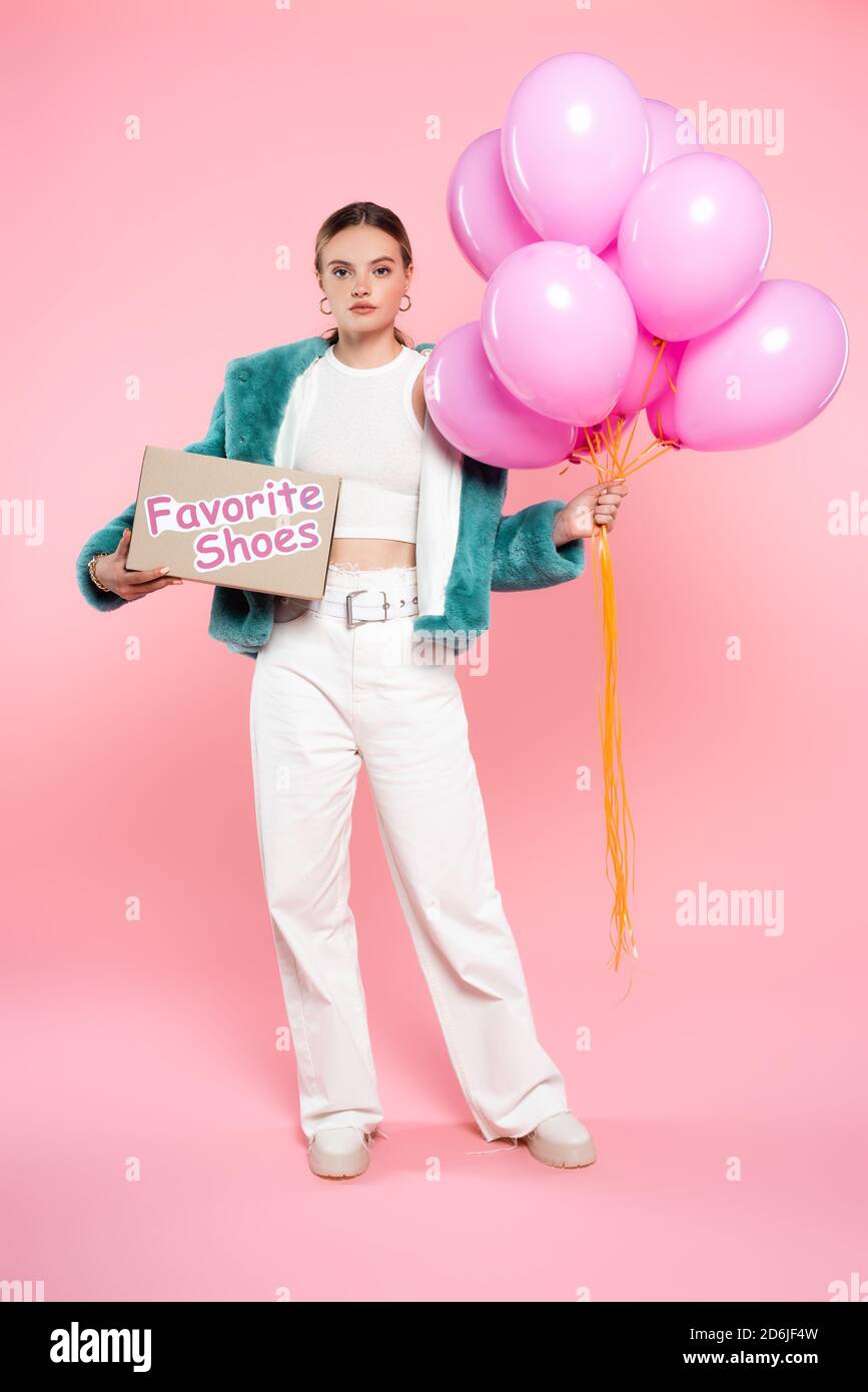 giovane donna che tiene scatola di cartone con le scritte preferite delle scarpe e. palloncini in piedi su rosa Foto Stock