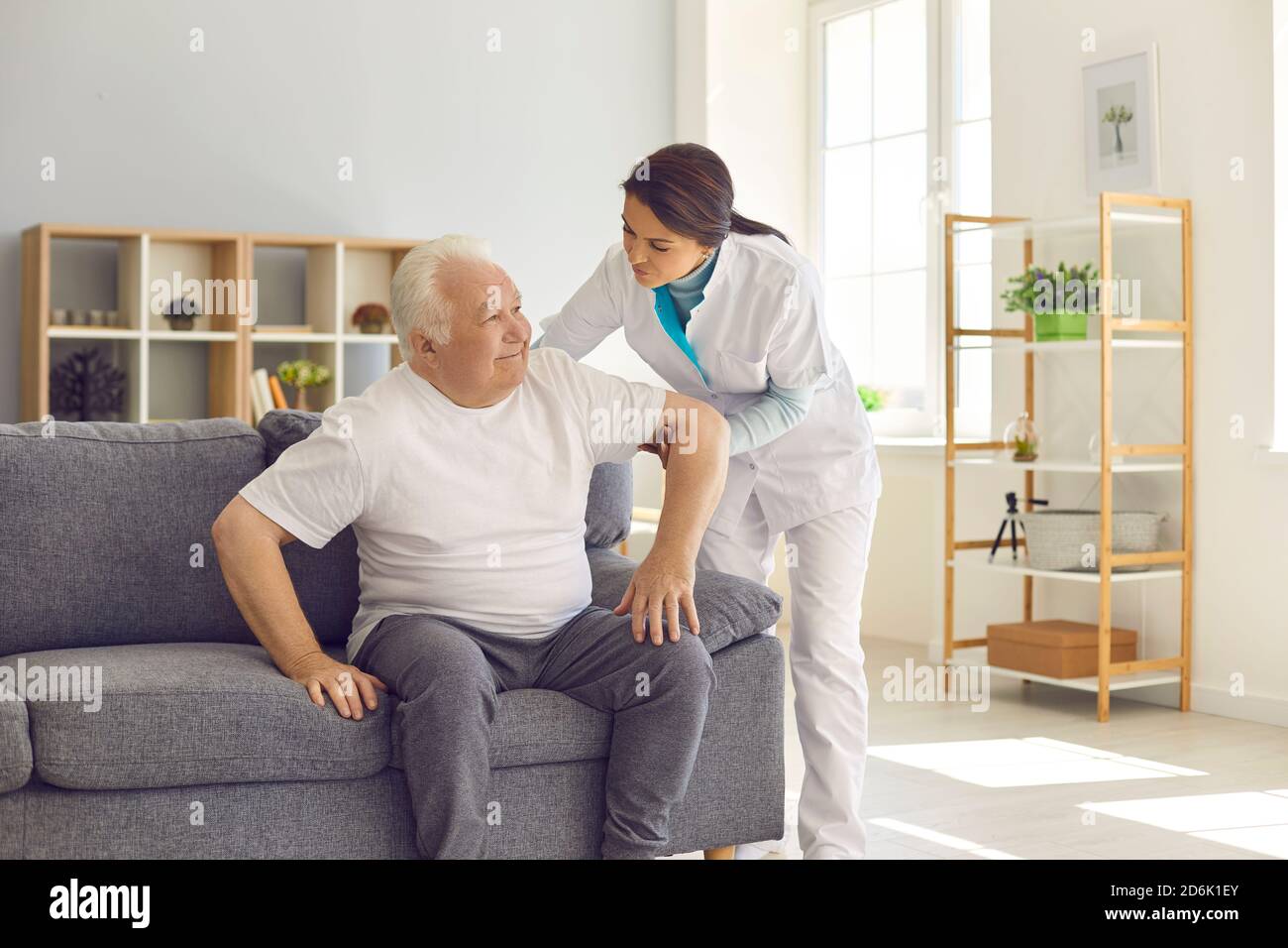 Infermiera che aiuta l'uomo anziano a levarsi in piedi in su dal divano dentro moderno ospedale o centro di riabilitazione geriatrica Foto Stock
