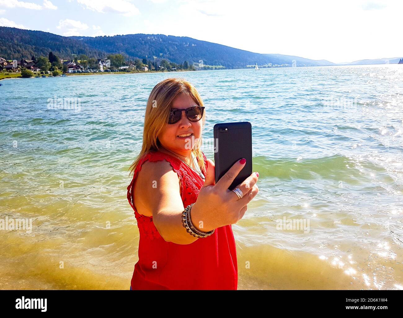 Donna in rosso che fa un selfie con il telefono cellulare sul bordo del lago Foto Stock