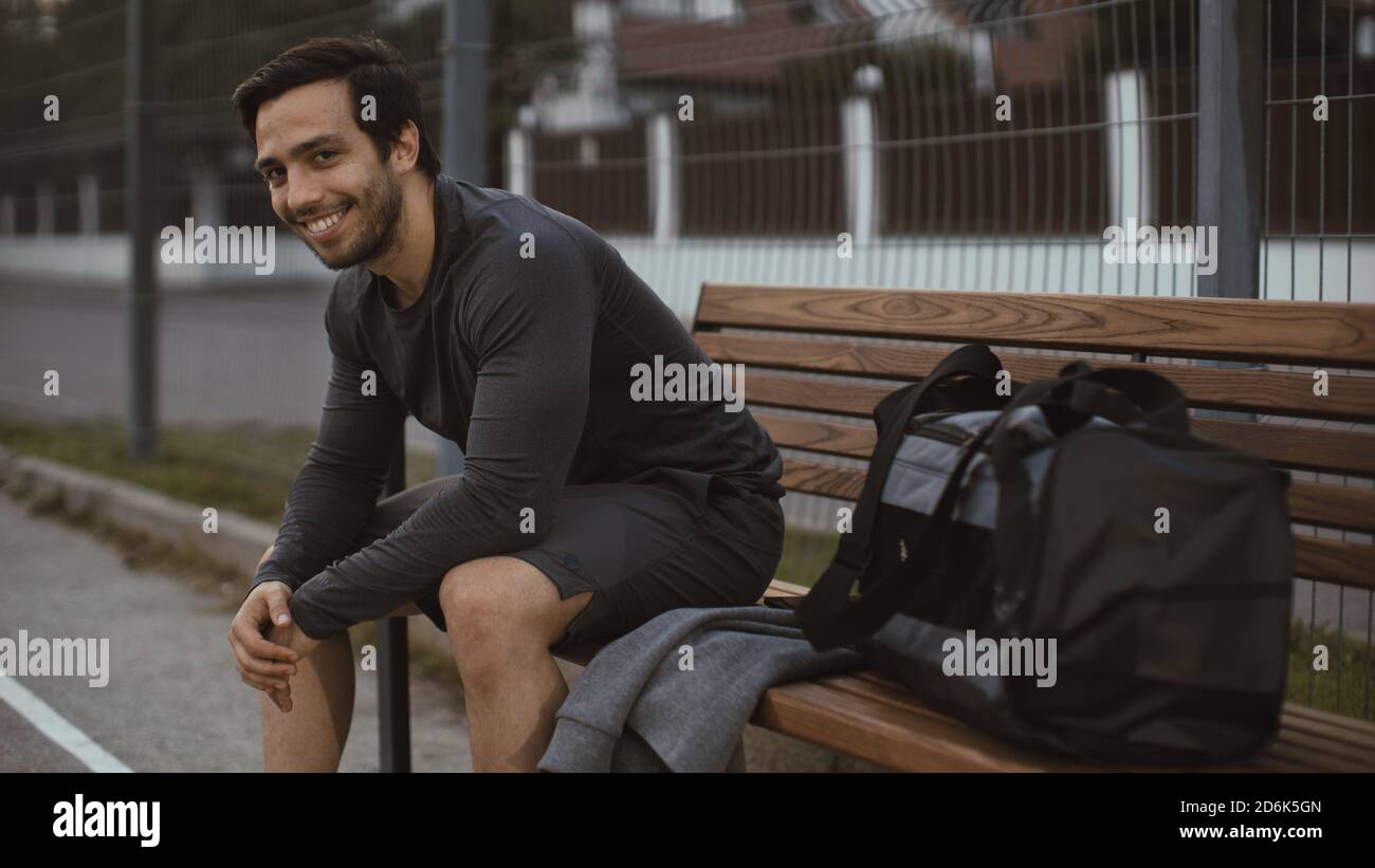L'atletico giovane uomo in outfit sportivo sta saltando su un banco e guardando nella macchina fotografica. Lui farà jogging in strada. Ambiente urbano. Foto Stock
