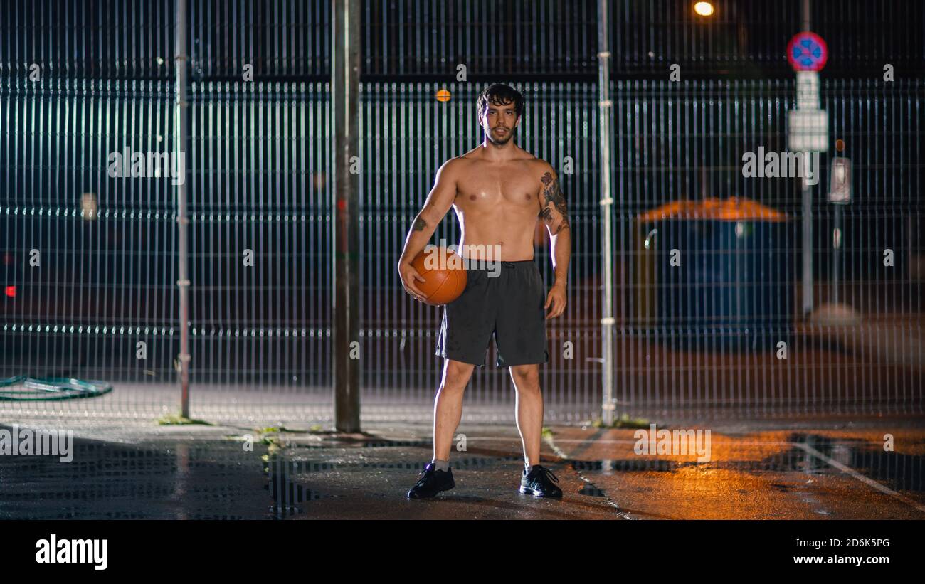 Atletico giovane maschio Basketball giocatore Sranging con la palla in posizione accovacciata in un quartiere residenziale recintato Streetball Court. Foto Stock