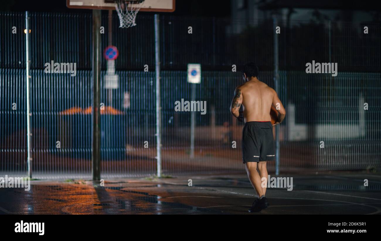 Atletico giovane maschio Basketball giocatore dribbles e getta la palla in posizione accovacciata in un quartiere residenziale recintato Streetball Court. Foto Stock