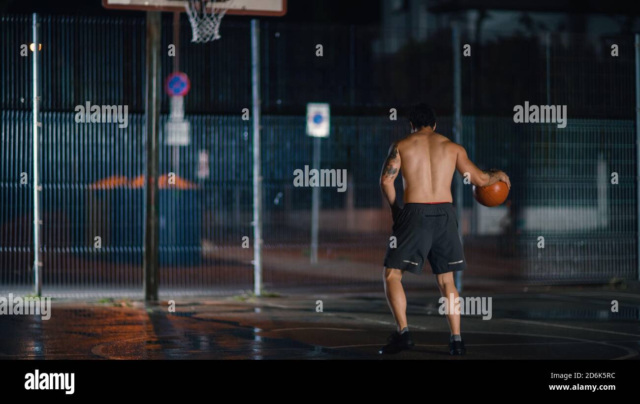 Atletico giovane maschio Basketball giocatore dribbles e getta la palla in posizione accovacciata in un quartiere residenziale recintato Streetball Court. Foto Stock