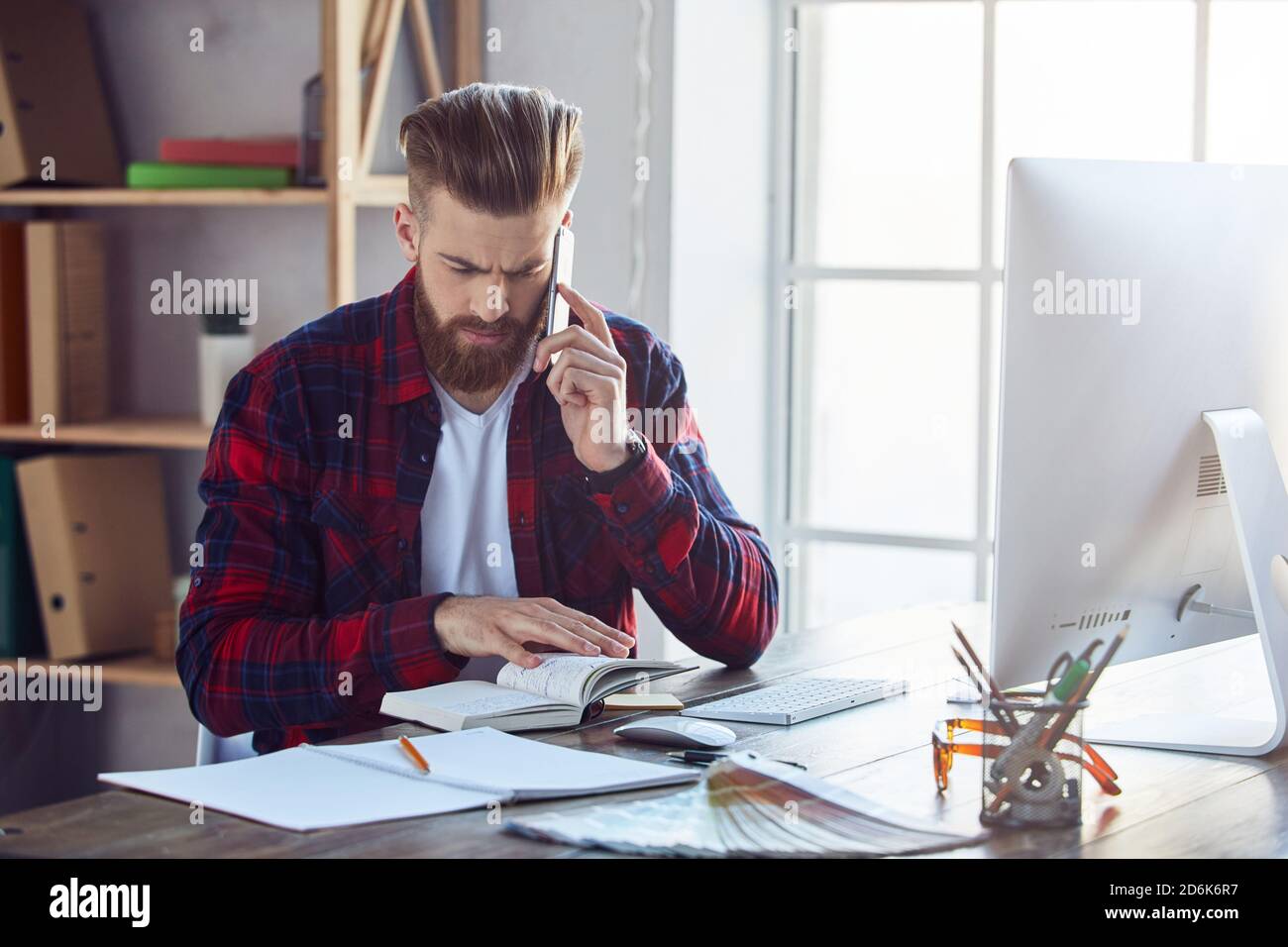 Uomo elegante con bearded che guarda in un notebook e parla al telefono mentre lavora in uno studio creativo. Concetto di stile di vita per l'ufficio Foto Stock