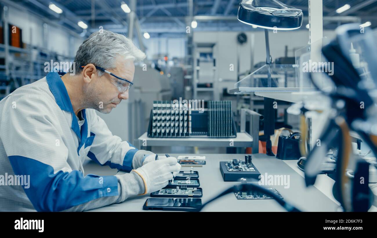 Senior Man in Blue - White Work Coat utilizza Plier per assemblare la scheda a circuito stampato per smartphone. Lavoratori di una fabbrica di elettronica in una High Tech Foto Stock