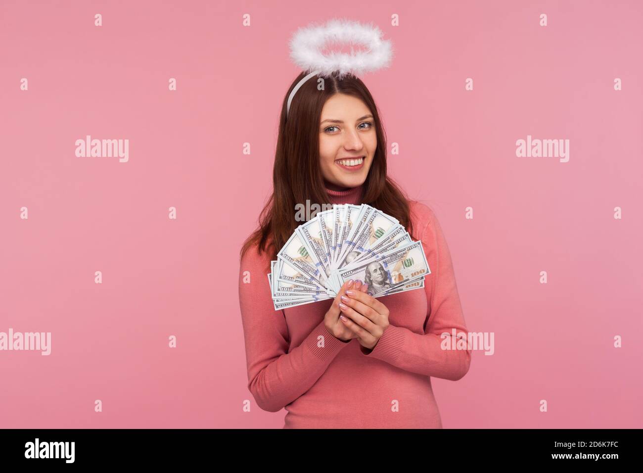 Donna angelica felice con alone sopra la testa tenendo e mostrando banconote in dollari, vantando grande vittoria della lotteria. Studio al coperto isolato su sfondo rosa Foto Stock