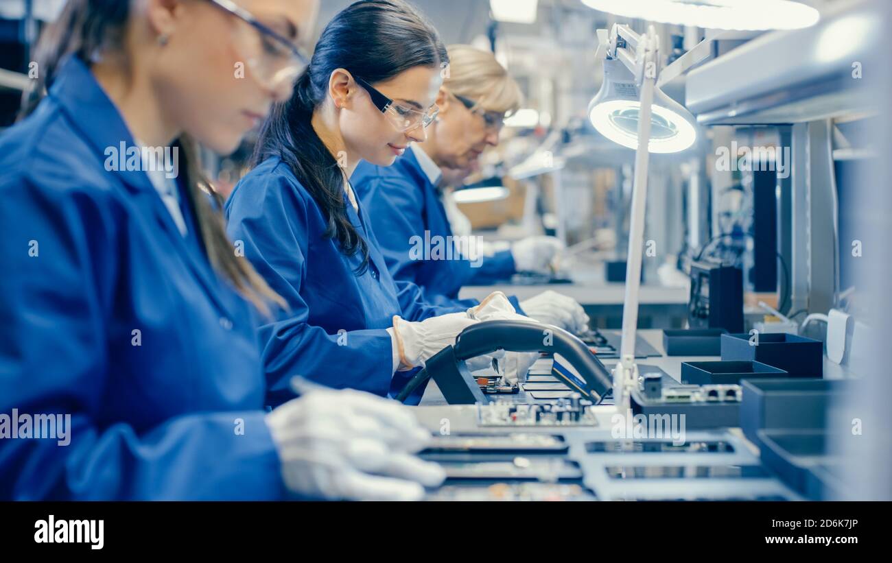 Lavoratori della Female Electronics Factory in Blue Work Coat e occhiali protettivi assemblando schede a circuito stampato per smartphone con pinzette. High Tech Foto Stock