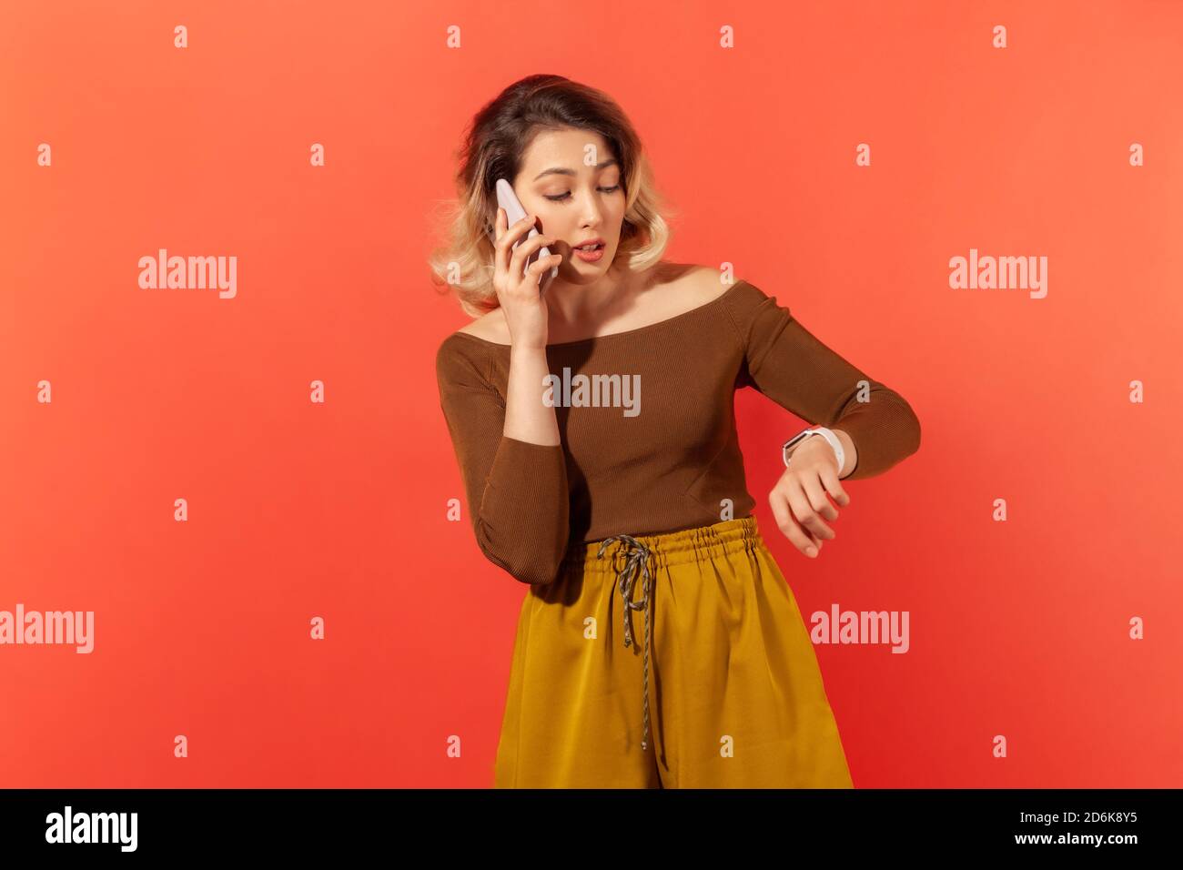 Bella donna che parla telefono guardando orologio sul polso, prendendo appuntamento o data. Studio al coperto isolato su sfondo arancione Foto Stock