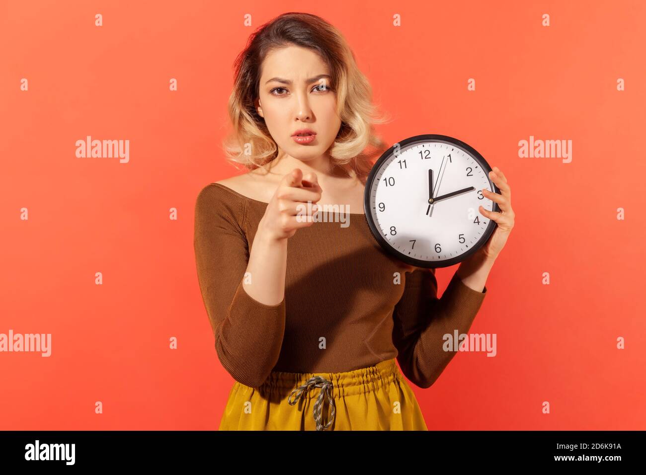 È ora di agire. Donna seria che tiene il grande orologio da parete che punta il dito a voi, motivazione, avvertimento circa la scadenza. Studio interno girato isolato su arancione ba Foto Stock