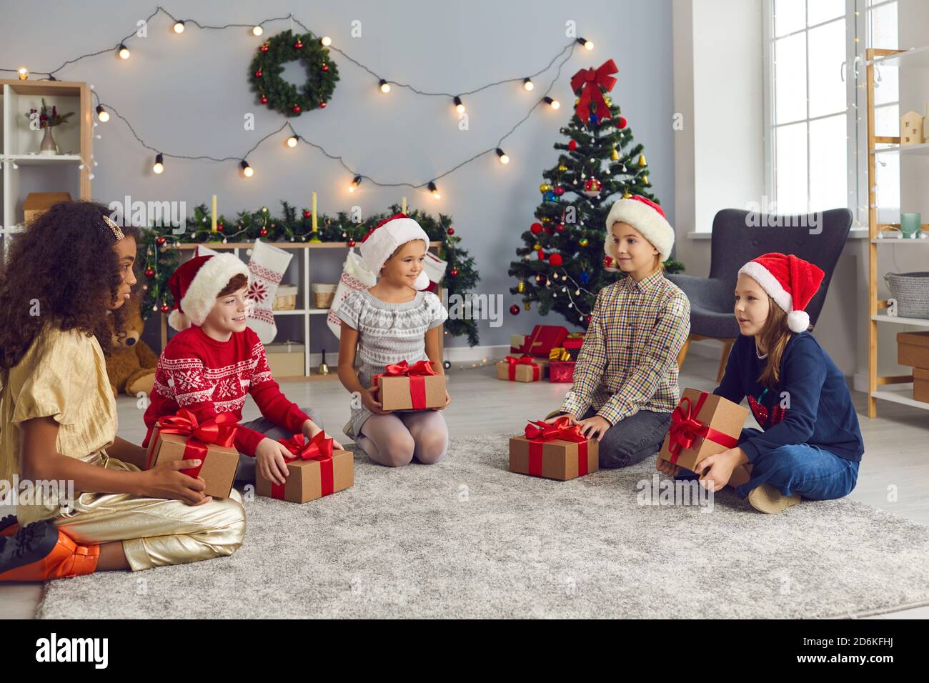 I bambini si siedono sul pavimento nel soggiorno la mattina di Natale, tenendo i loro regali e parlando Foto Stock