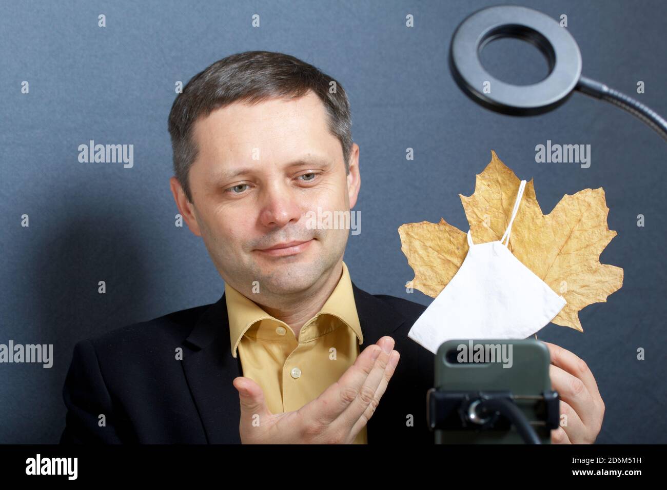 Un uomo in costume senza cravatta. Dimostra una foglia di acero essiccata su una maschera medica. Davanti a lui c'è un supporto per uno smartphone con una lampada per l'illumina Foto Stock