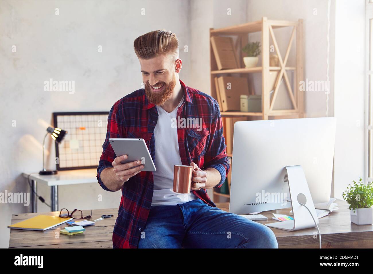 Sorridente giovane ed elegante uomo che guarda lo schermo dei gadget mentre ti godi caffè durante la pausa nel suo studio creativo Foto Stock