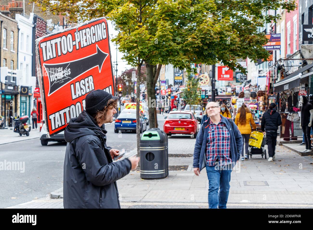 Un uomo che tiene un cartello che fa pubblicità al tatuaggio di Nemesis e che piercing su Camden High Street, Londra, Regno Unito Foto Stock