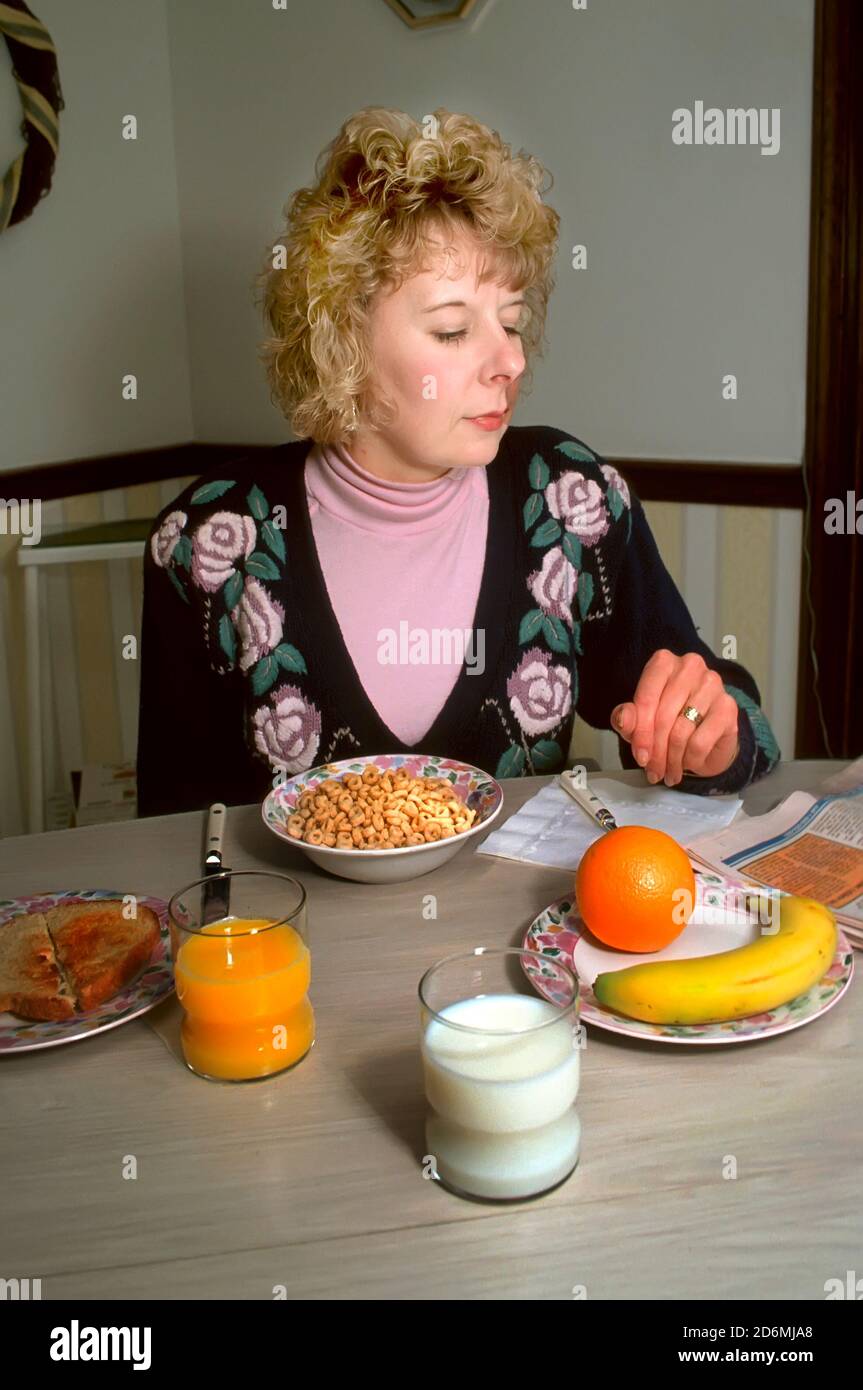 La donna adulta mangia una sana colazione che comprende cereali, succo di frutta, latte e frutta mentre legge il giornale Foto Stock