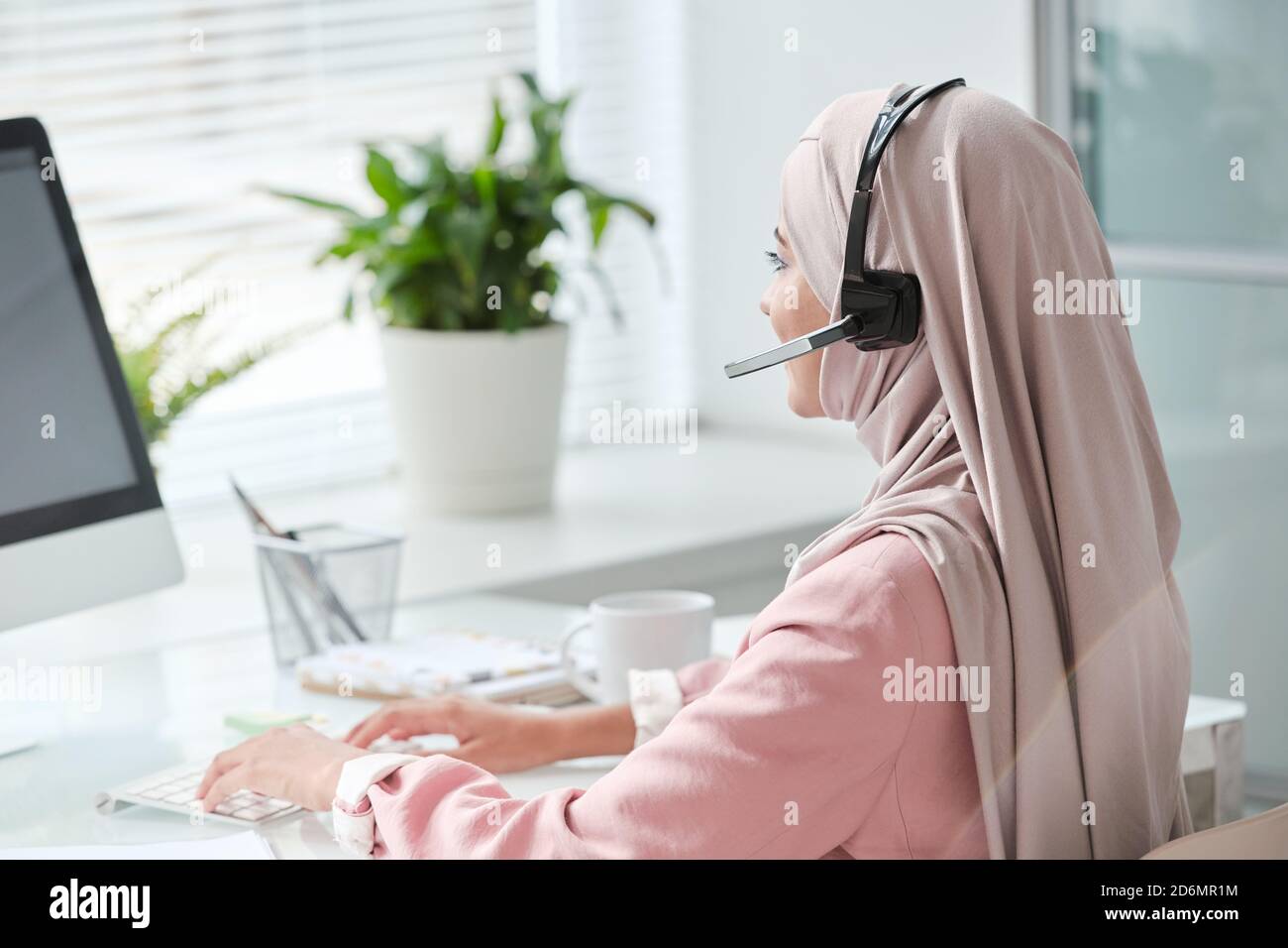 Giovane operatore musulmano contemporaneo in hijab e cuffie dattilografa in ufficio Foto Stock