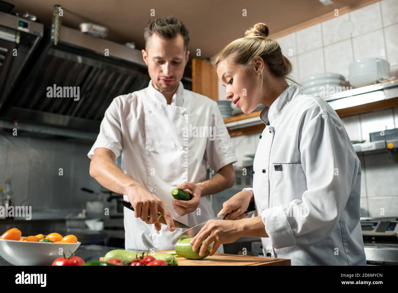 Giovane chef professionista sicuro con coltello e cetriolo che lo consultano tirocinante Foto Stock