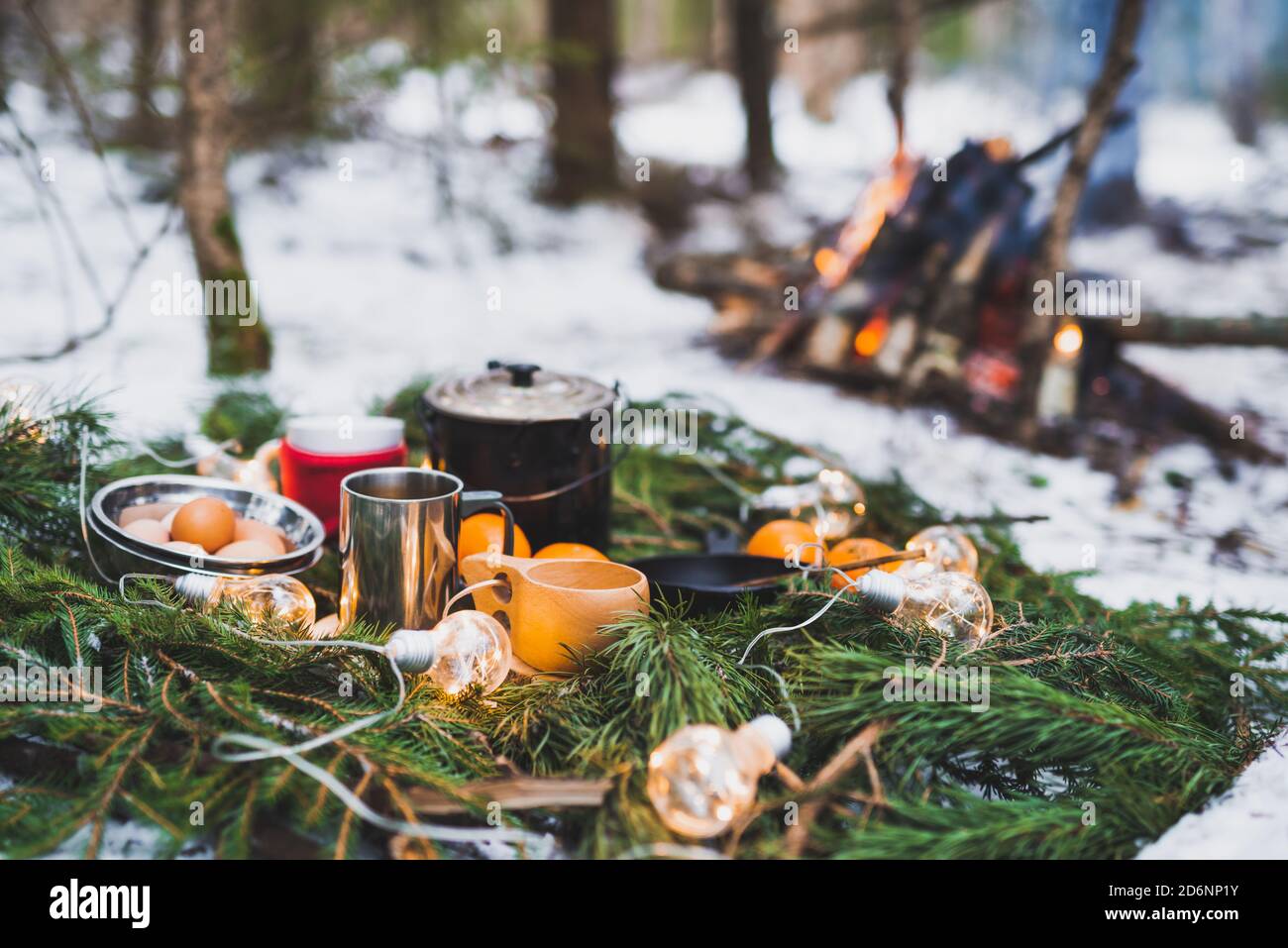 Picnic invernale nella neve vicino al fuoco con arance e tè. Ghirlande di Natale su rami di abete Foto Stock
