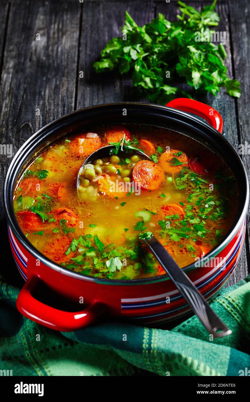 Zuppa di piselli spezzati con salsiccia bavarese, pastinaca, carote con prezzemolo e cipolla verde servita su una pentola rossa antiaderente con un mestolo di zuppa su una da Foto Stock