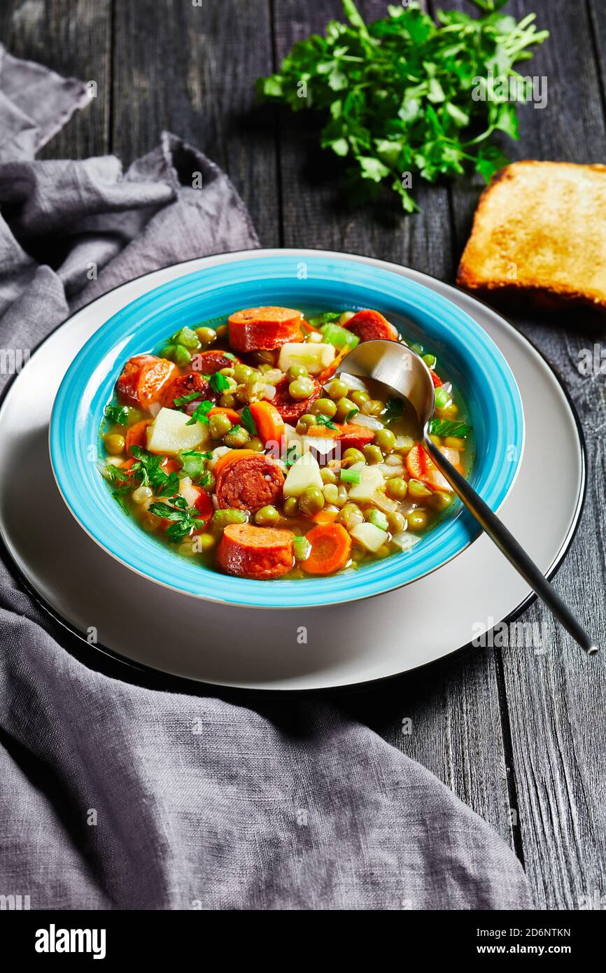 Zuppa tedesca di piselli spezzati con salsiccia bavarese, pastinaca, carote condite con prezzemolo e scalone servita su un piatto blu con un cucchiaio su un bac di legno scuro Foto Stock