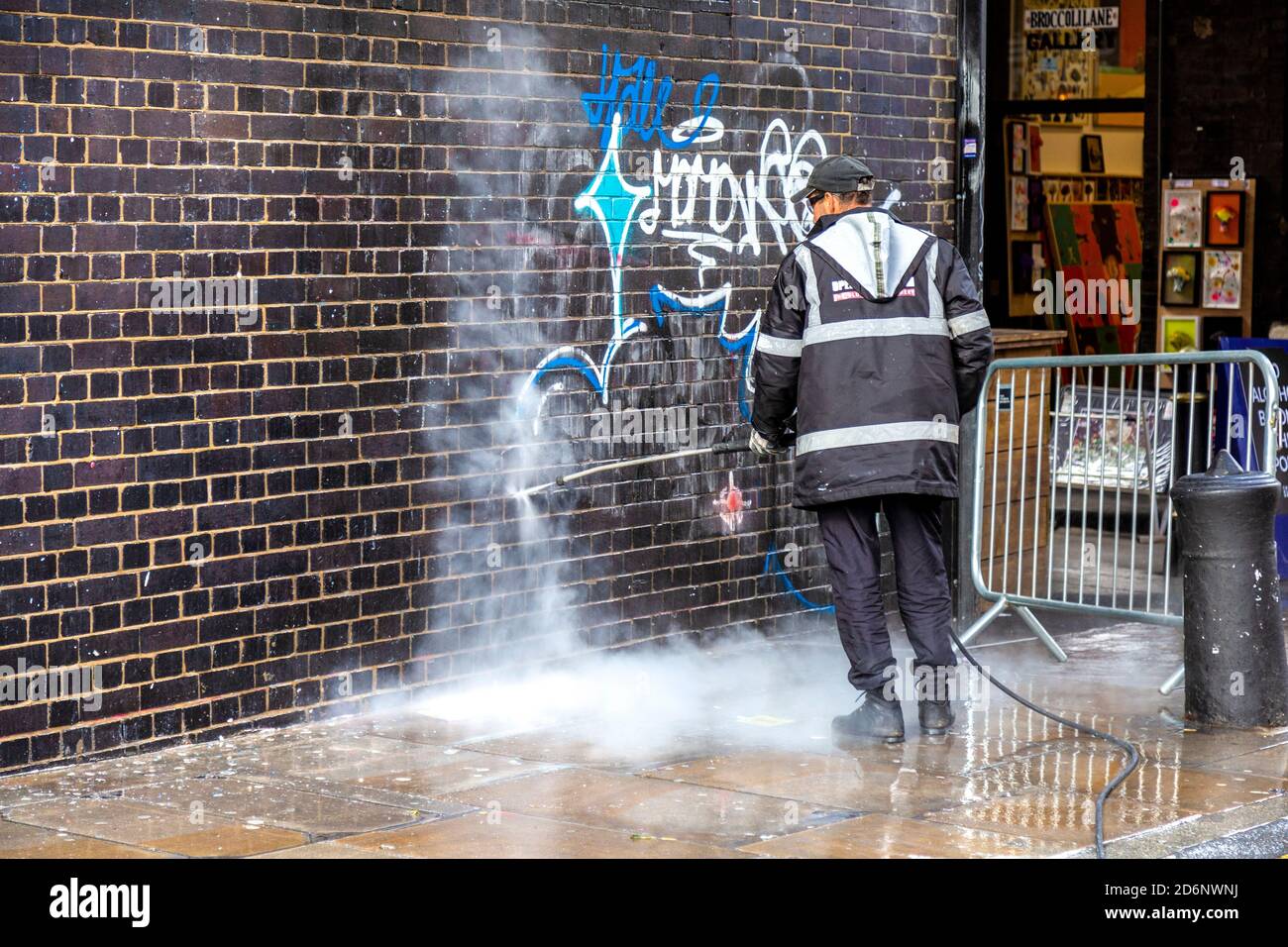 Uomo che pulisce graffiti da un muro di mattoni con un lavatore a pressione su Brick Lane, East London, UK Foto Stock