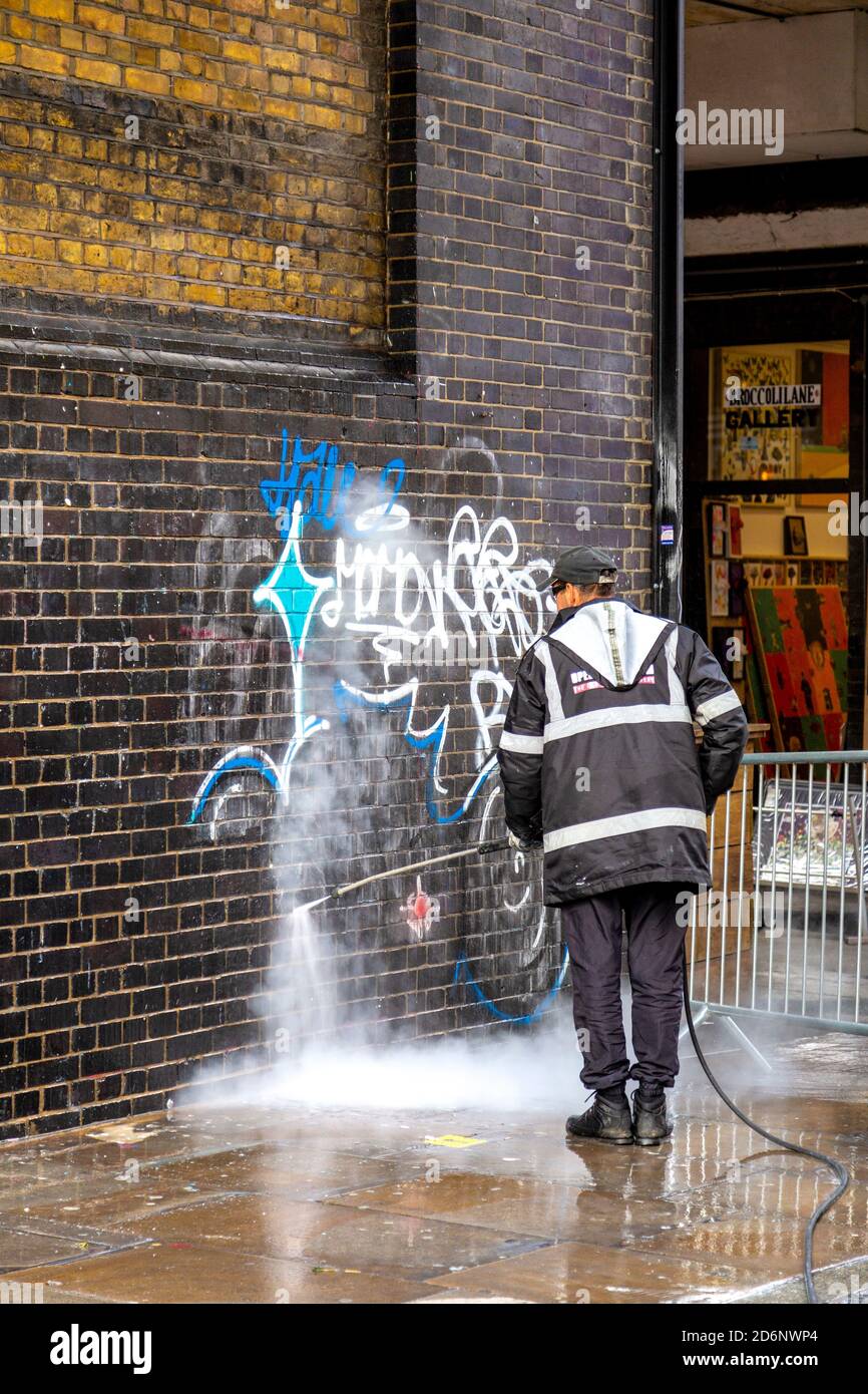 Uomo che pulisce graffiti da un muro di mattoni con un lavatore a pressione su Brick Lane, East London, UK Foto Stock