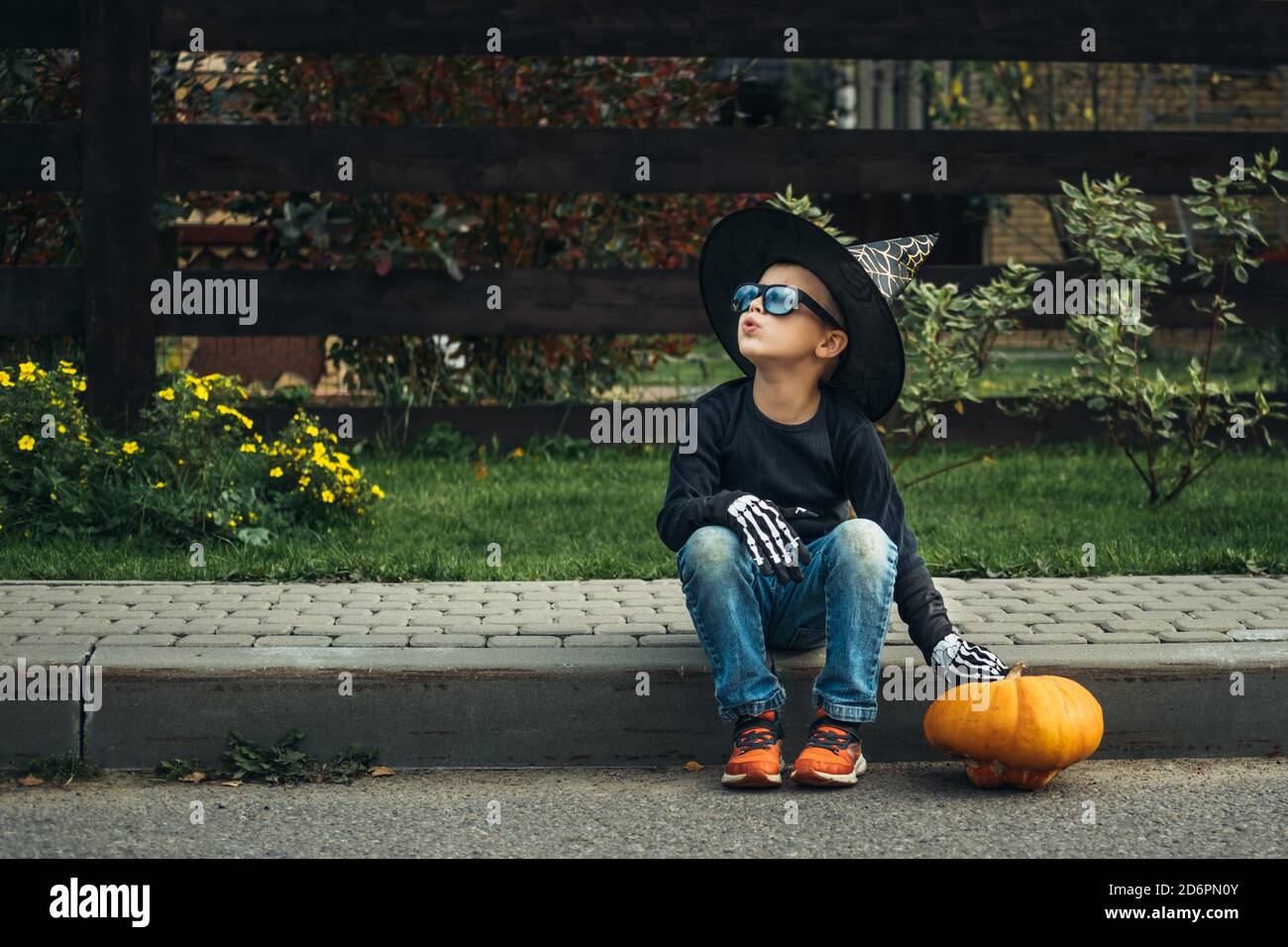 Il ragazzo in cappello e guanti da costume, e occhiali da sole con zucca seduta sulla strada. Halloween felice. Halloween bambini. Foto Stock