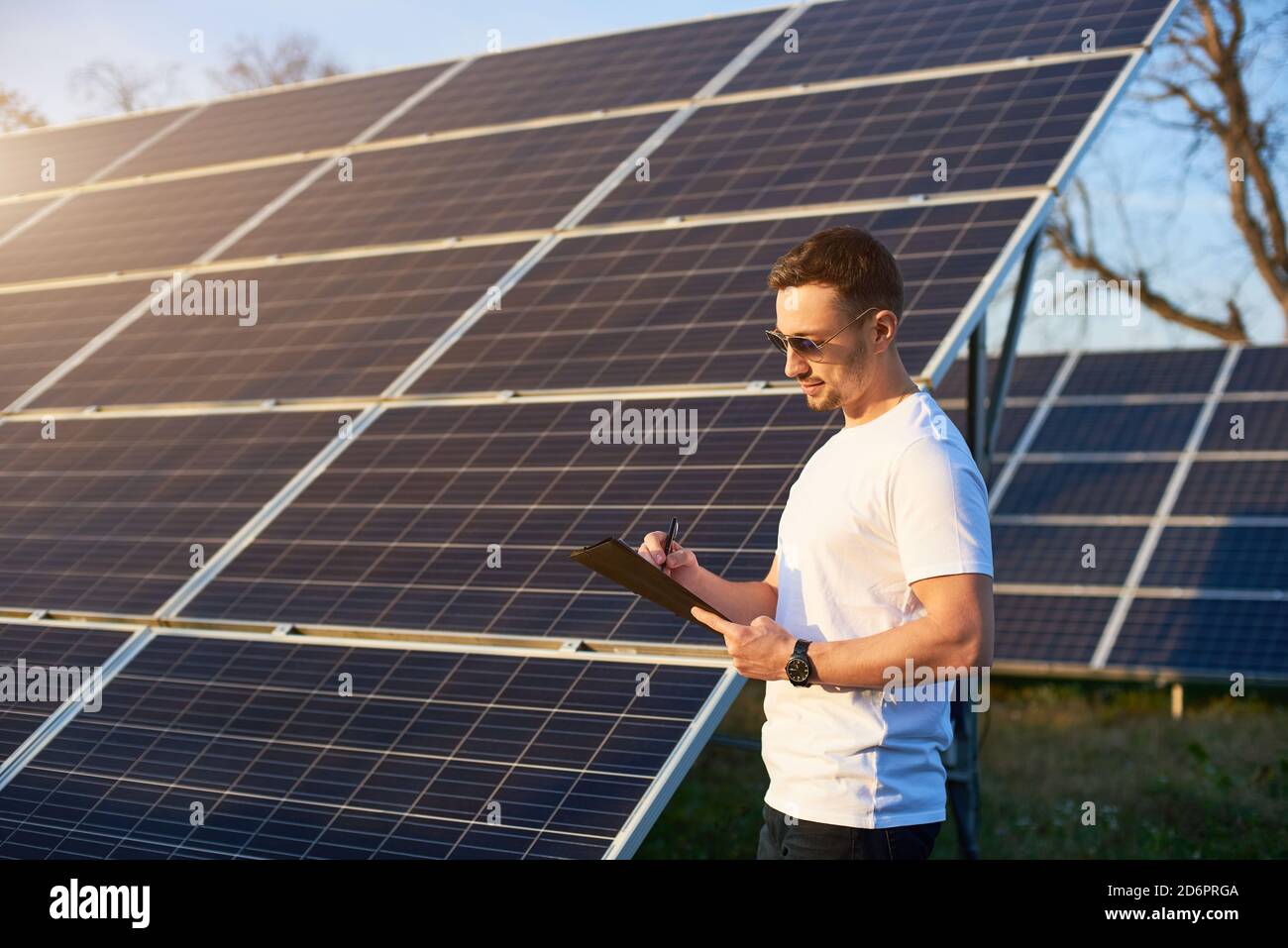 Ritratto di un giovane uomo di successo in piedi da un impianto fotovoltaico solare che guarda nella sua cartella meditando su un certo piano di affari e prendendo appunti, vista laterale, concetto di energia alternativa Foto Stock