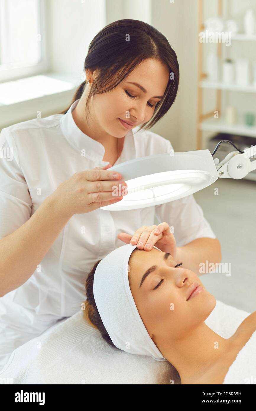 Donna dermatologa che esamina l'elasticità e la condizione facciali del womans sotto lampada Foto Stock